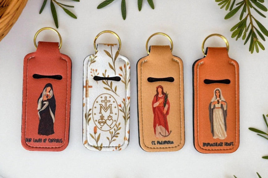 catholic chapstick keychains with catholic illustrations on a marble surface with greenery.